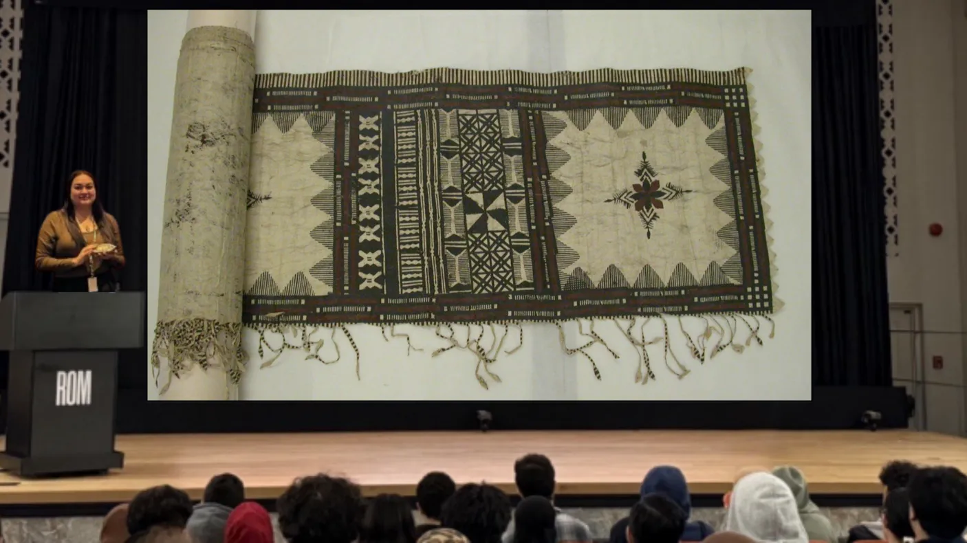 Une femme indigène souriante se tient derrière un podium sur une scène. Sur un écran à côté d'elle, on voit l'image d'un morceau de tissu avec des motifs complexes à l'encre noire et brune.