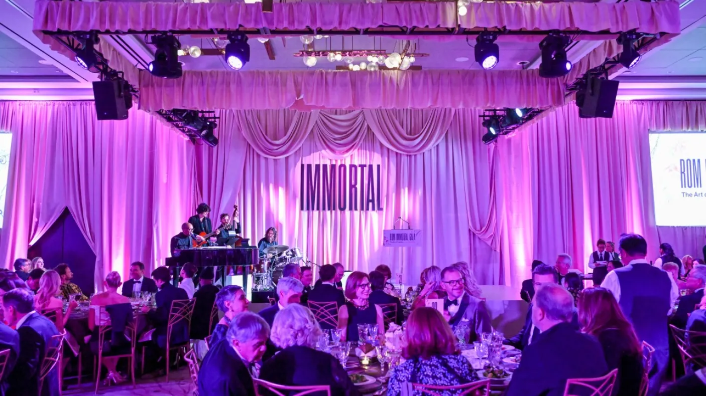 A banquet hall with round tables filled with people under purple lighting and a stage with musicians playing.