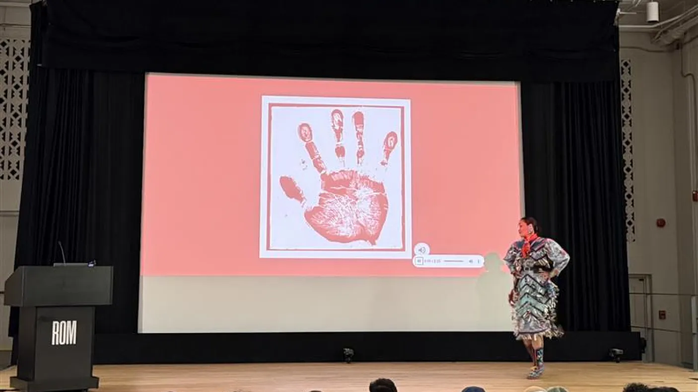 Une femme autochtone portant une robe en jingle se tient sur la scène d'un théâtre. L'écran derrière elle montre une empreinte de main blanche sur un écran rouge.
