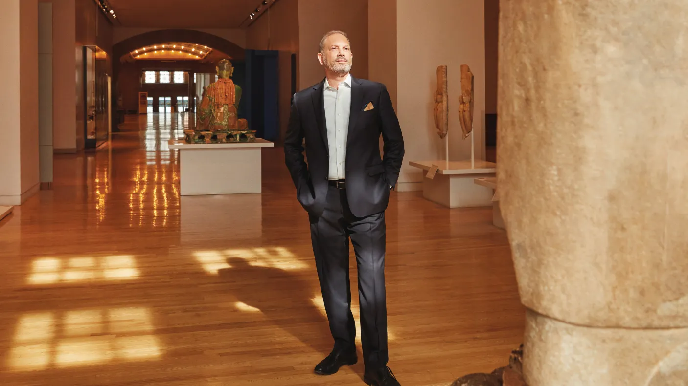 A man in a black tuxedo standing in a spacious gallery with wooden floors, stone columns, and display cases.