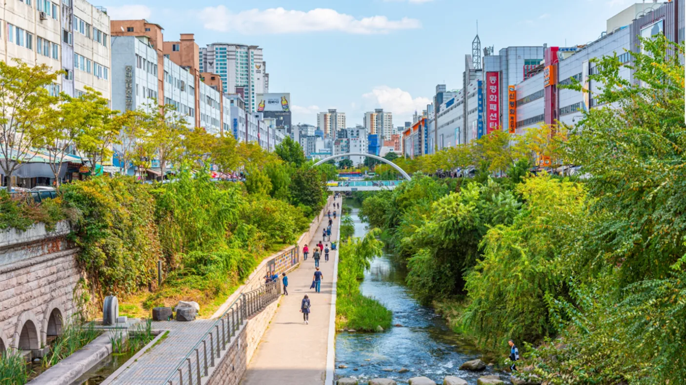 Urban park with a walking path, railway tracks on the left, trees, and a river flowing through the middle under a blue sky.