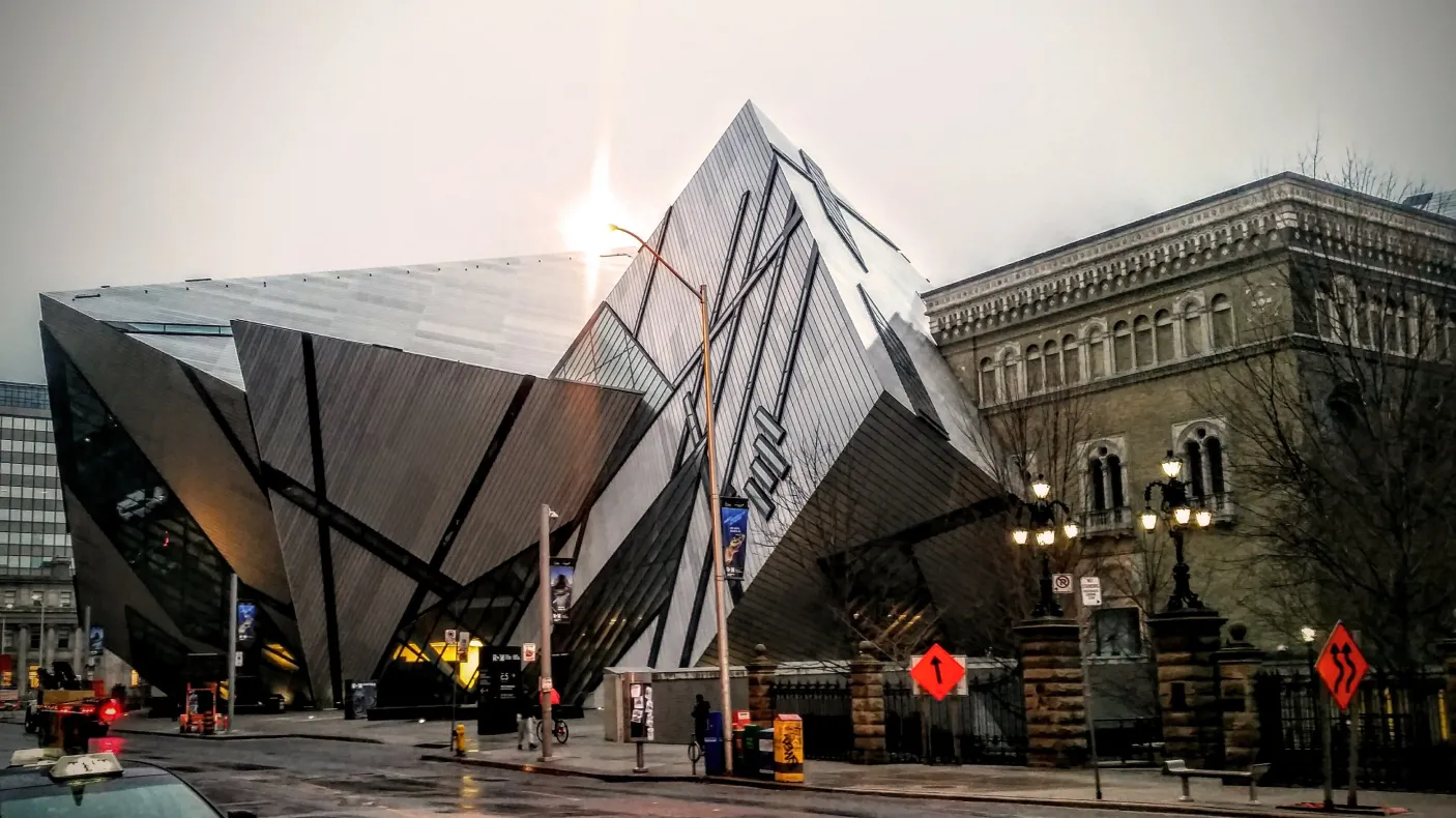 Modern angular glass and metal building facade of the Royal Ontario Museum with sunlight behind it, and a historic building in the background.