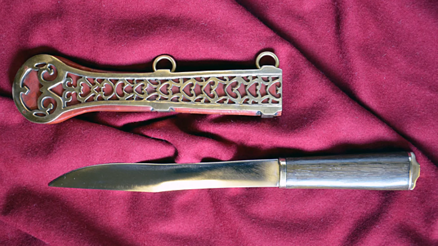 Steel knife with a straight blade and a polished metal handle next to an ornately decorated metal sheath on red cloth.