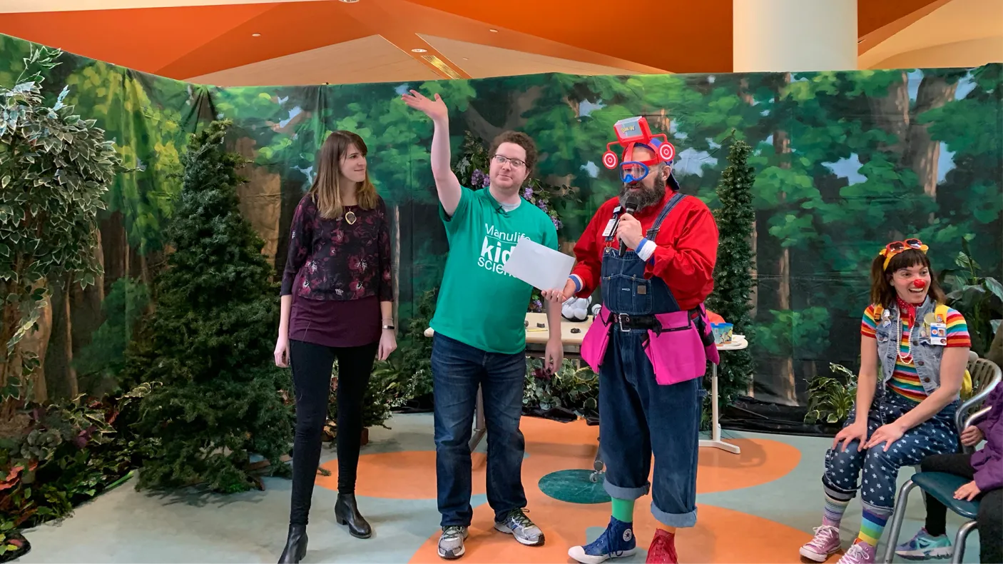 Three people stand on a colorful stage with a leafy green backdrop, one person in a red costume holds an item, and one person is seated on the right.