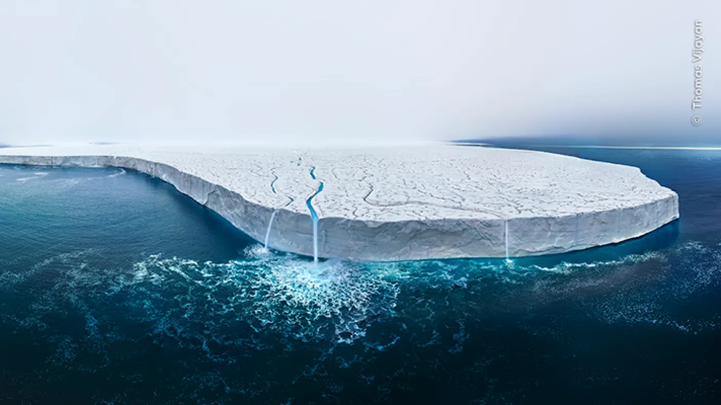 A large ice shelf floating on calm, dark blue ocean water under a clear sky.