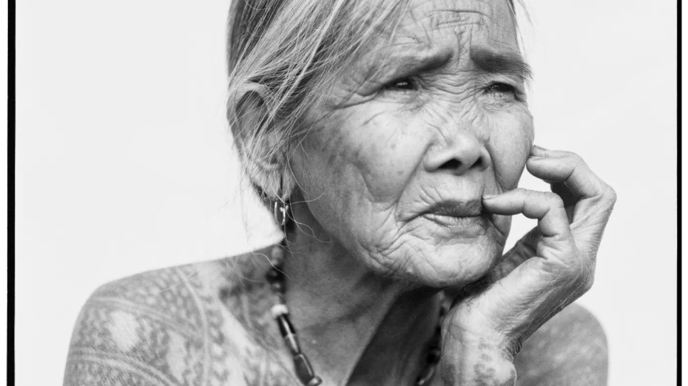 Elderly woman with traditional tattoos on her arms and upper chest, resting her chin on her hand.