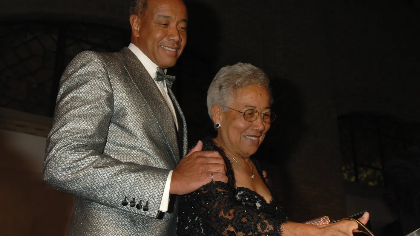 A man in a silver suit jacket stands behind a woman in glasses and a black lace dress.
