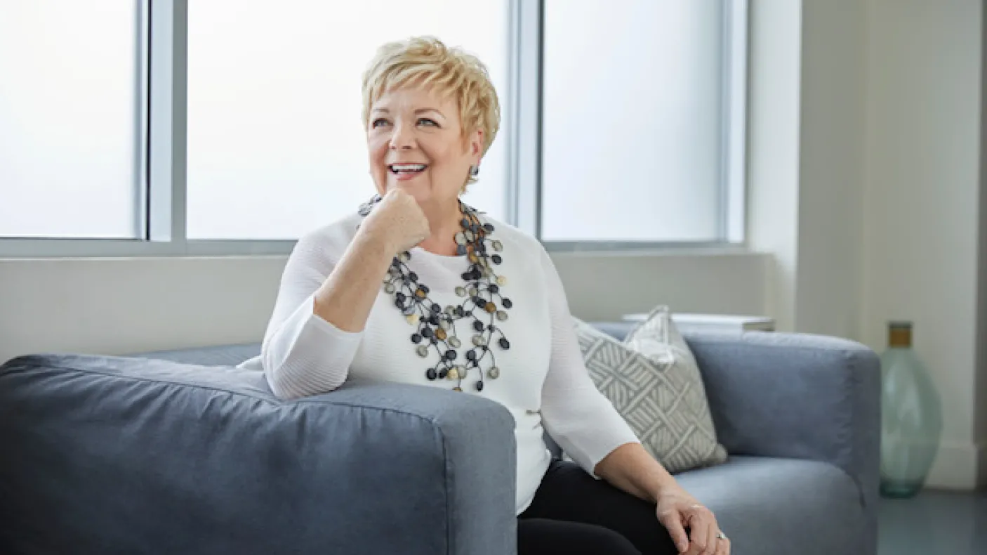 Portrait of Janice Price smiling and seated on a gray sofa, wearing a white blouse and a statement necklace.