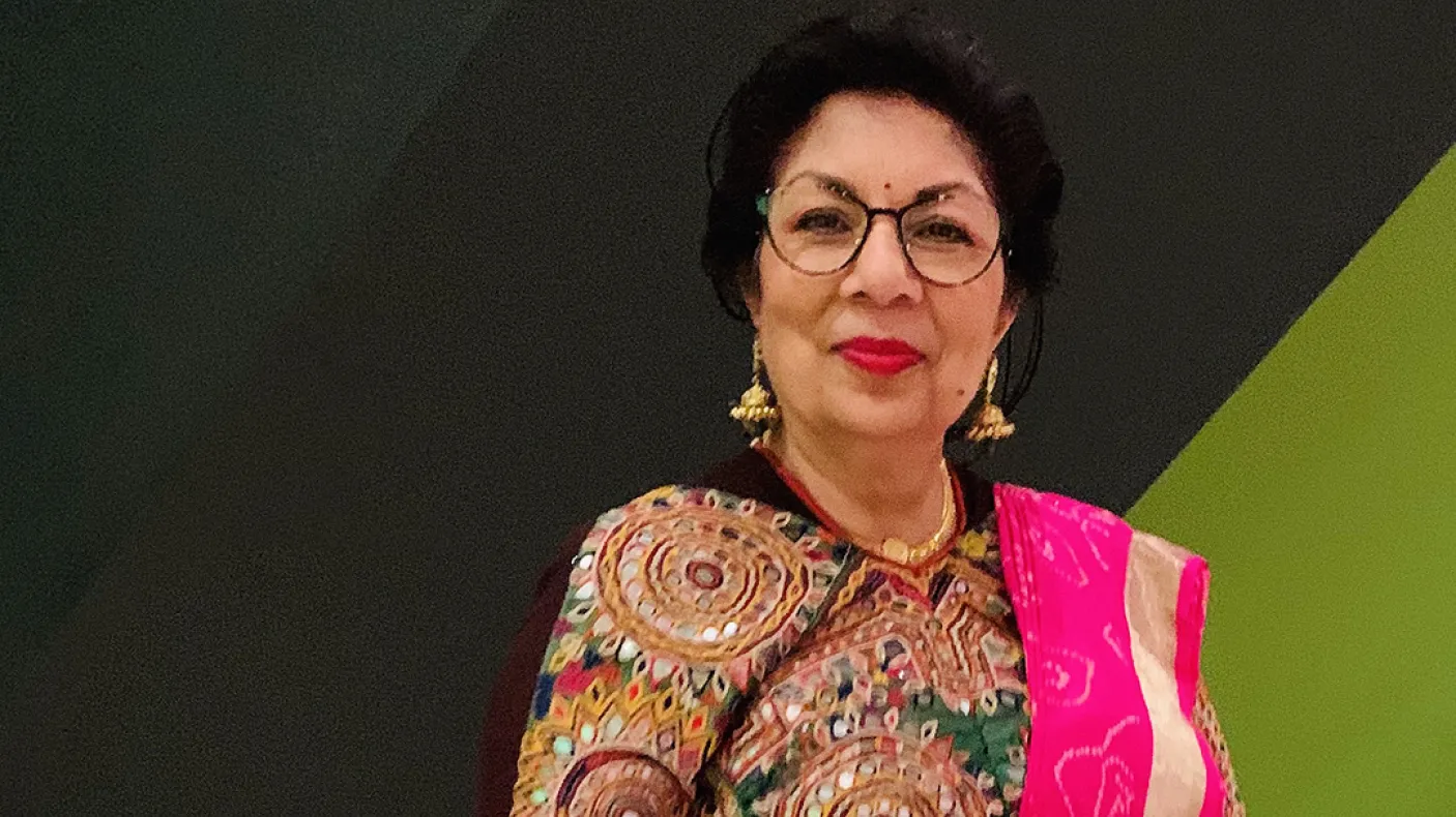 A woman wearing a brown patterned top, gold necklace, glasses, and a bright pink shawl on her left shoulder against a green and black background.
