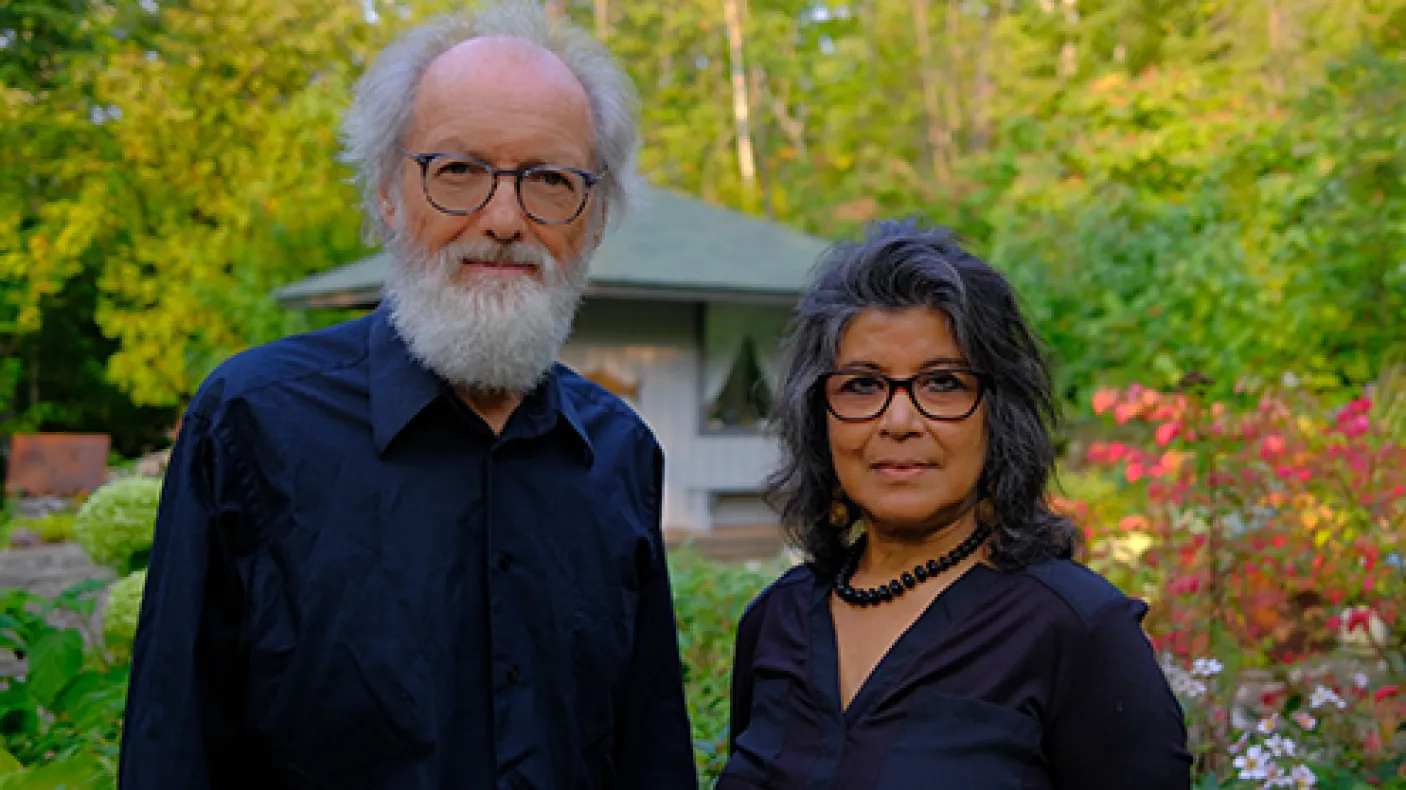 Portrait of an older man with white hair and beard wearing glasses and a black shirt, and a middle-aged woman with dark hair and glasses wearing a black top, standing outdoors with green trees and a pavilion in the background.