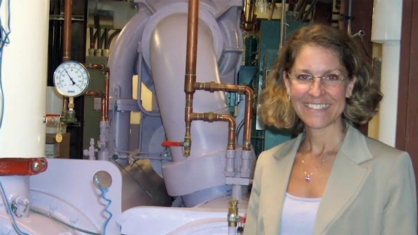 A woman with glasses and curly hair stands smiling in front of industrial pipes and gauges.