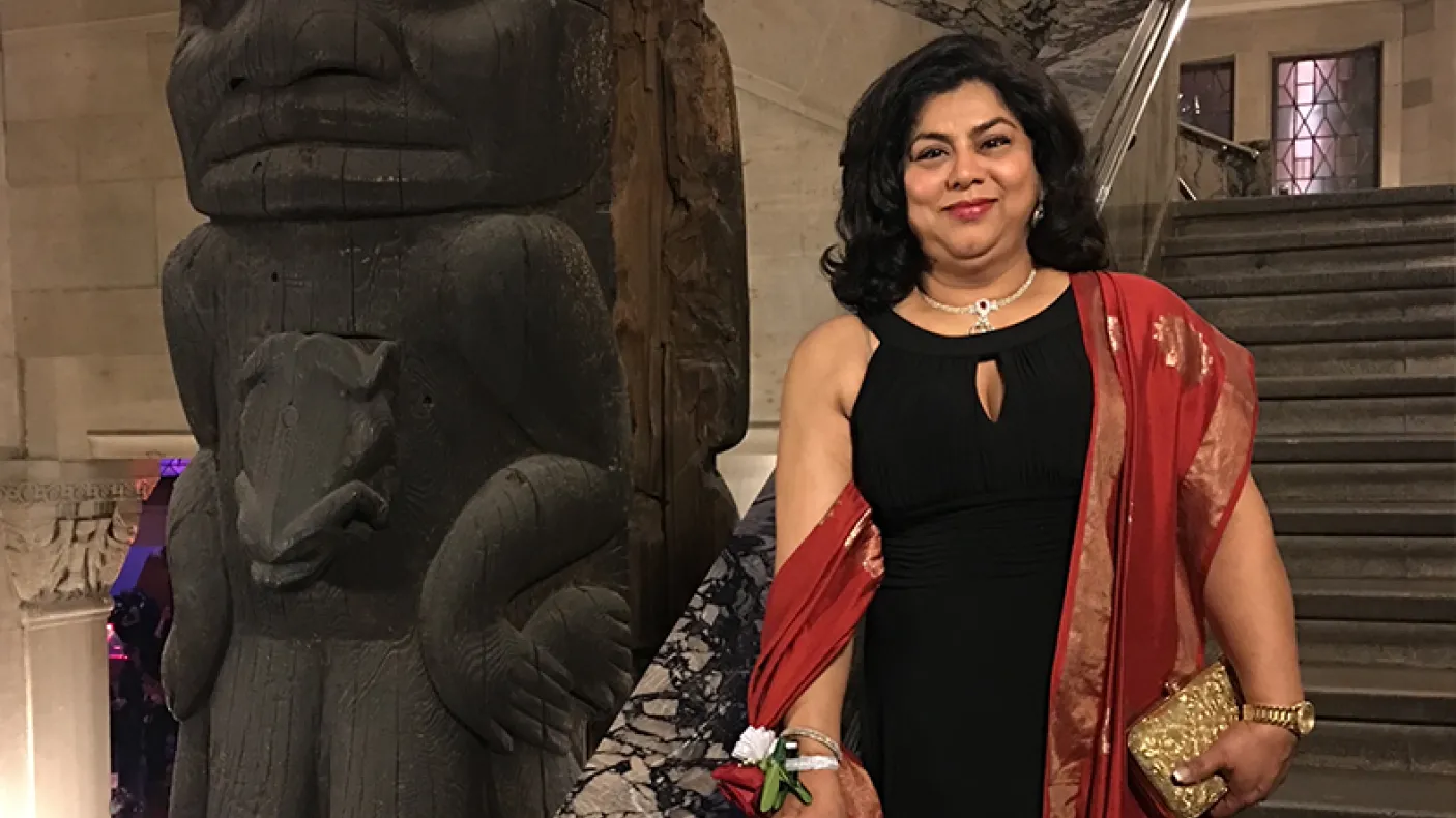 A woman in a black dress with a red shawl stands beside a large carved wooden totem pole inside a building.