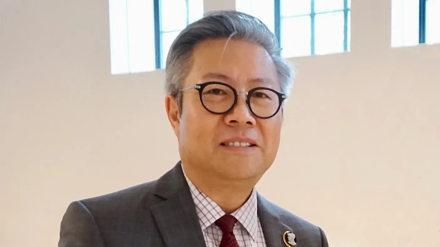 Man with glasses wearing a dark suit and red patterned tie, standing indoors against a beige wall.