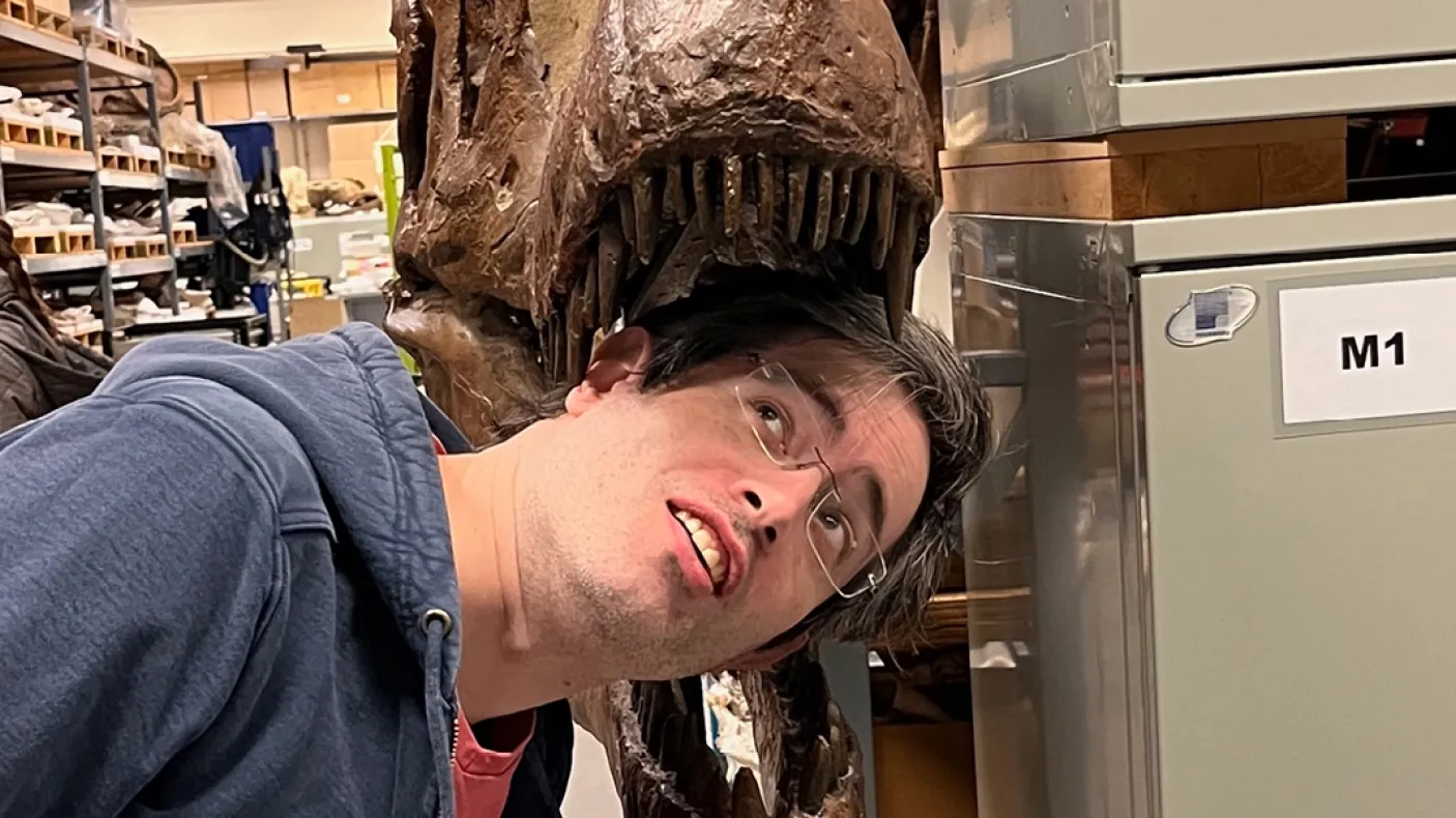 A person with dark hair leans their head toward the jaws of a large dinosaur skull mounted on a display stand.