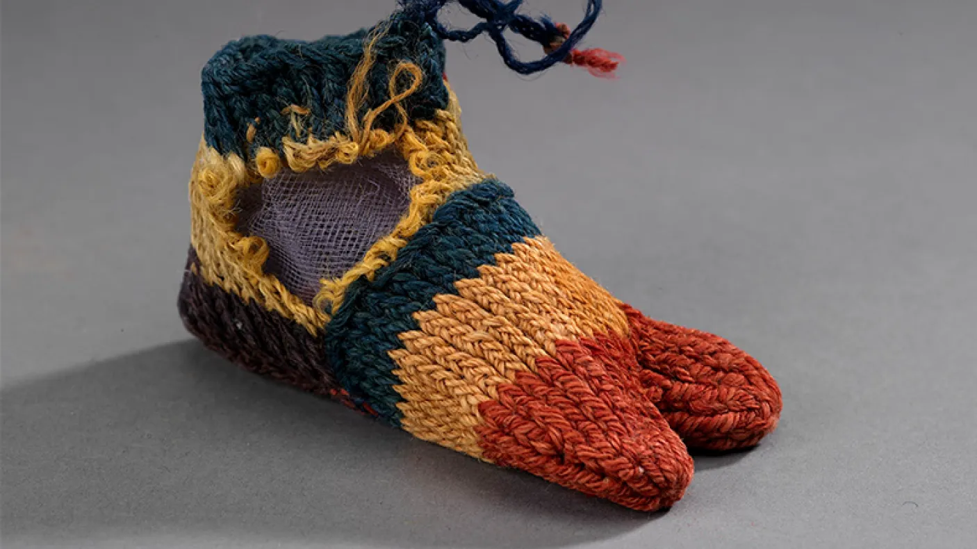 Woven baby shoes with orange toes, yellow and green bands, and blue laces on gray background.