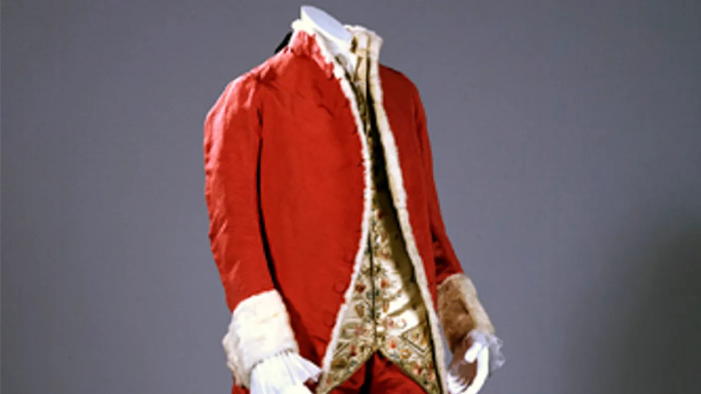 Red 18th-century coat with white lace cuffs and a patterned waistcoat underneath.