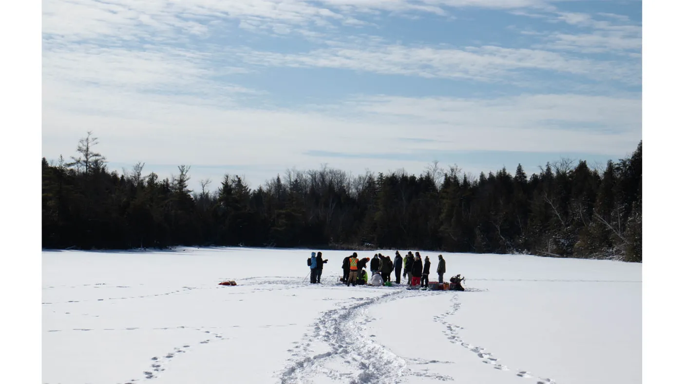 Group of people gathered on a snowy frozen lake surrounded by trees under a partly cloudy sky.