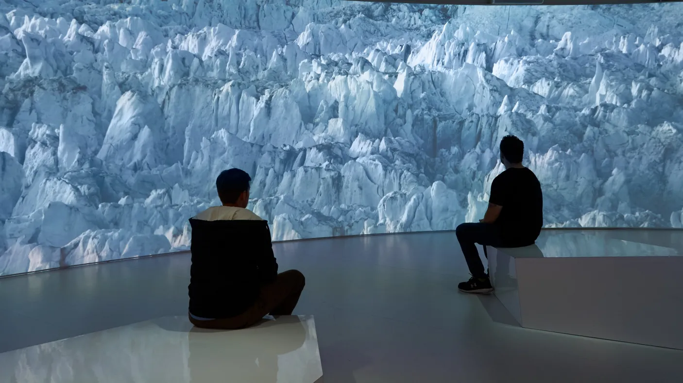 Two people sit on benches facing a large digital wall displaying a blue-hued glacier scene.