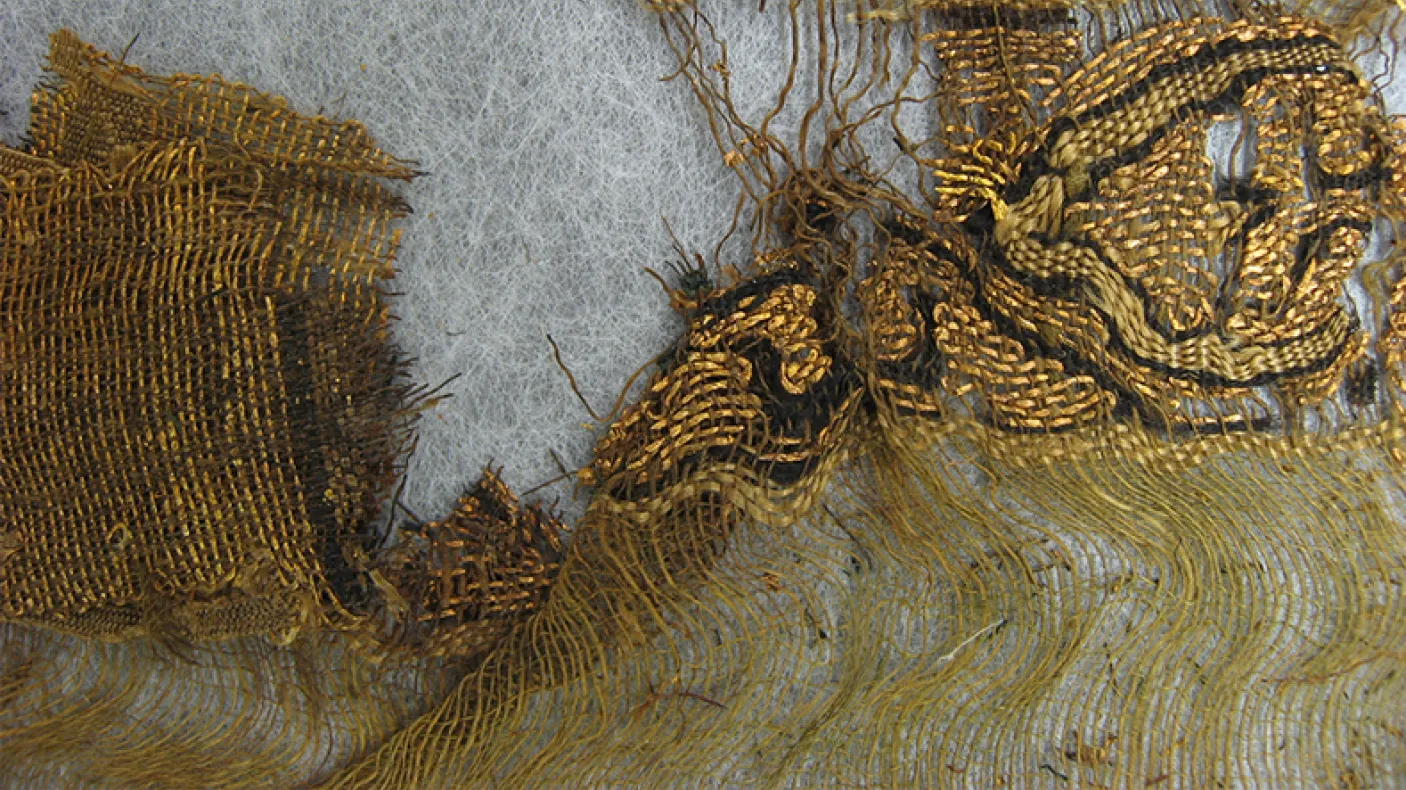 Fragment of ancient textile showing loose threads and woven fabric in brown and beige tones.