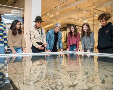 Seven people are gathered around and looking at a large glass-covered exhibit table with a map or drawing underneath.