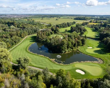Aerial view of a golf course hole with a pond, sand bunkers, trees, and expansive green fields under a partly cloudy sky.