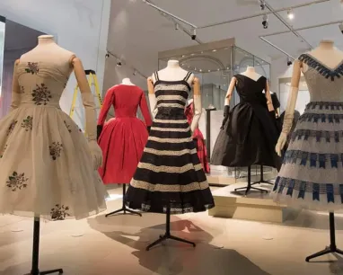 Seven vintage-style dresses displayed on mannequins in a museum exhibit with a lighted floor and walls.