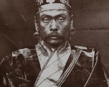 Portrait of a man in traditional attire, wearing a headpiece and a patterned sash across his chest.
