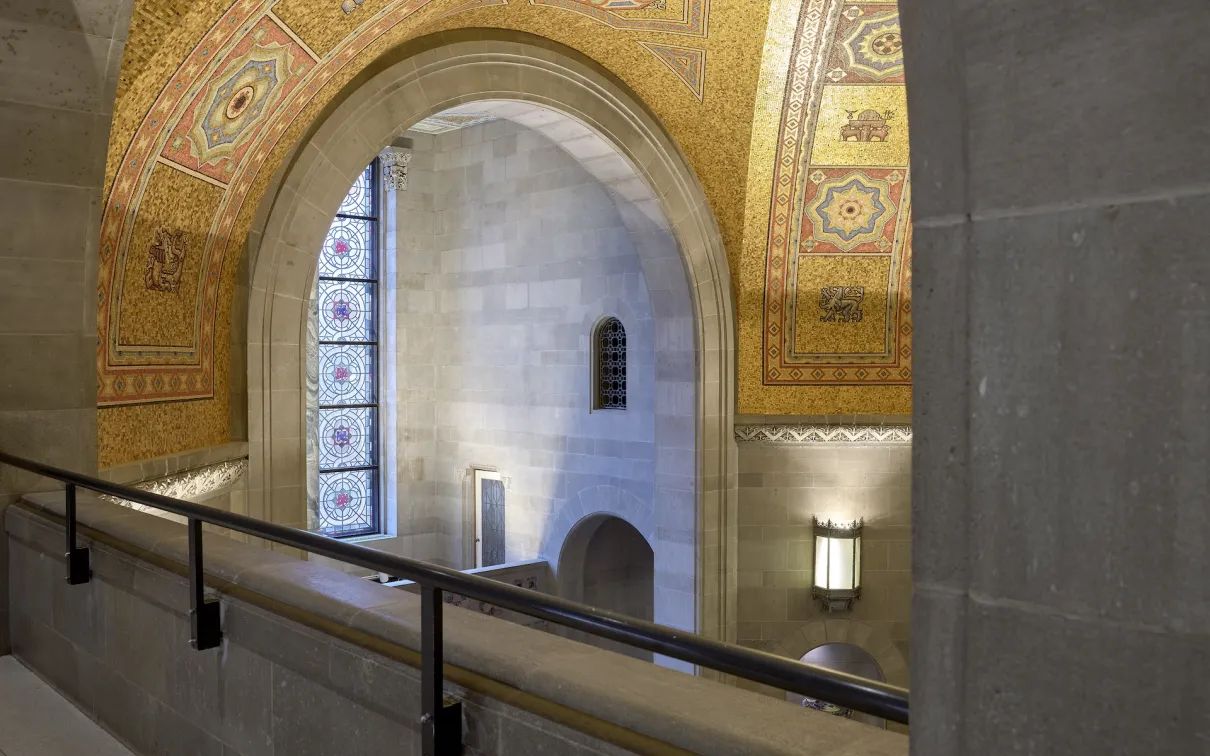 Stone interior arches and walls with a mosaic gold-patterned arch above and a metal railing in front.