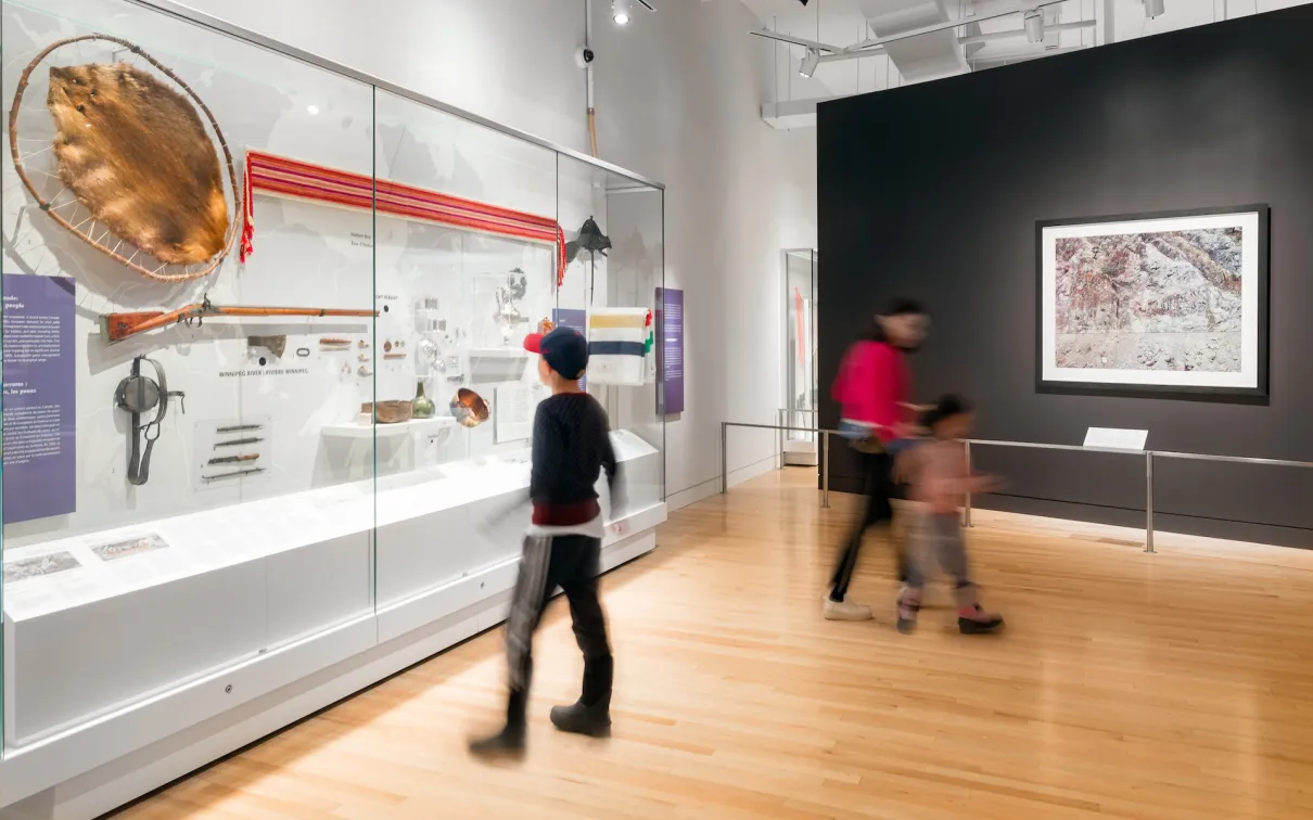 Museum gallery with glass display cases containing artifacts and two visitors walking by.
