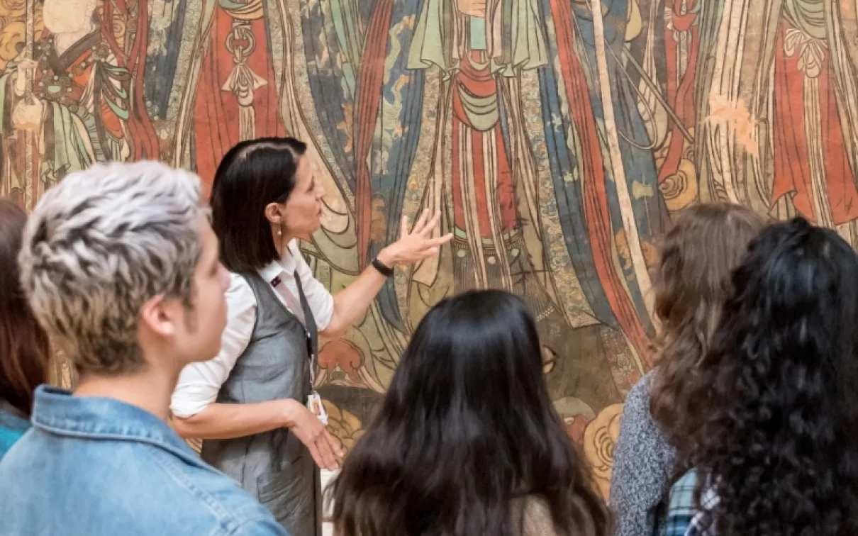 A group of four people view and discuss a large, colorful mural or painting featuring figures in traditional robes.