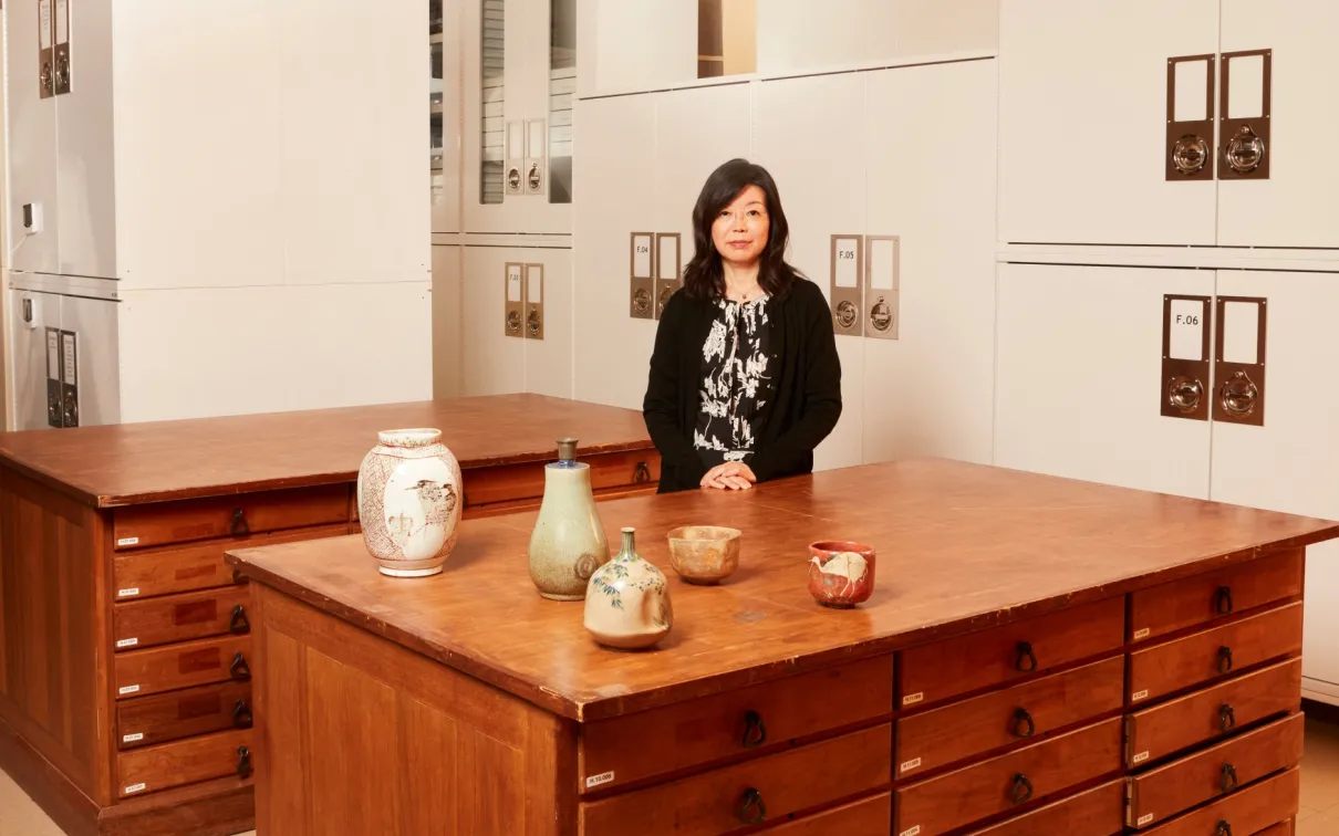 A woman stands behind a wooden cabinet with drawers, displaying five ceramic vessels of varying shapes and sizes.