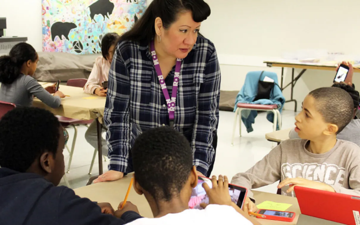 A teacher in a plaid shirt engages with four students seated around a table in a classroom.