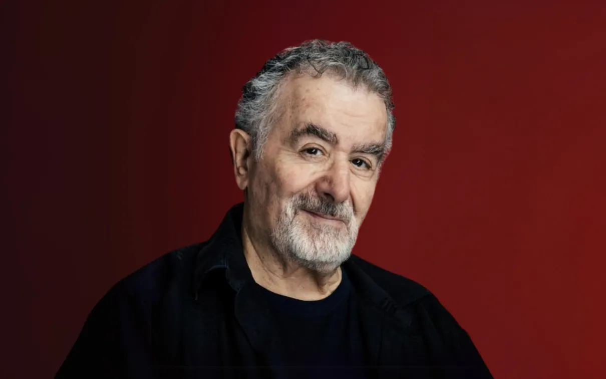 Portrait of an older man with gray hair and beard wearing a black shirt against a maroon background.
