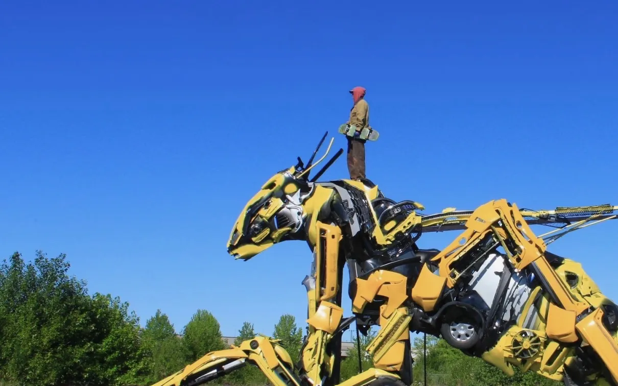 Large yellow mechanical dinosaur sculpture made from industrial parts on a wooden platform outdoors under a clear blue sky.