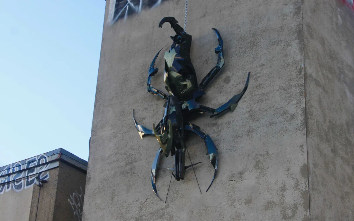 Large blue and black metal beetle sculpture mounted on a beige concrete building wall.