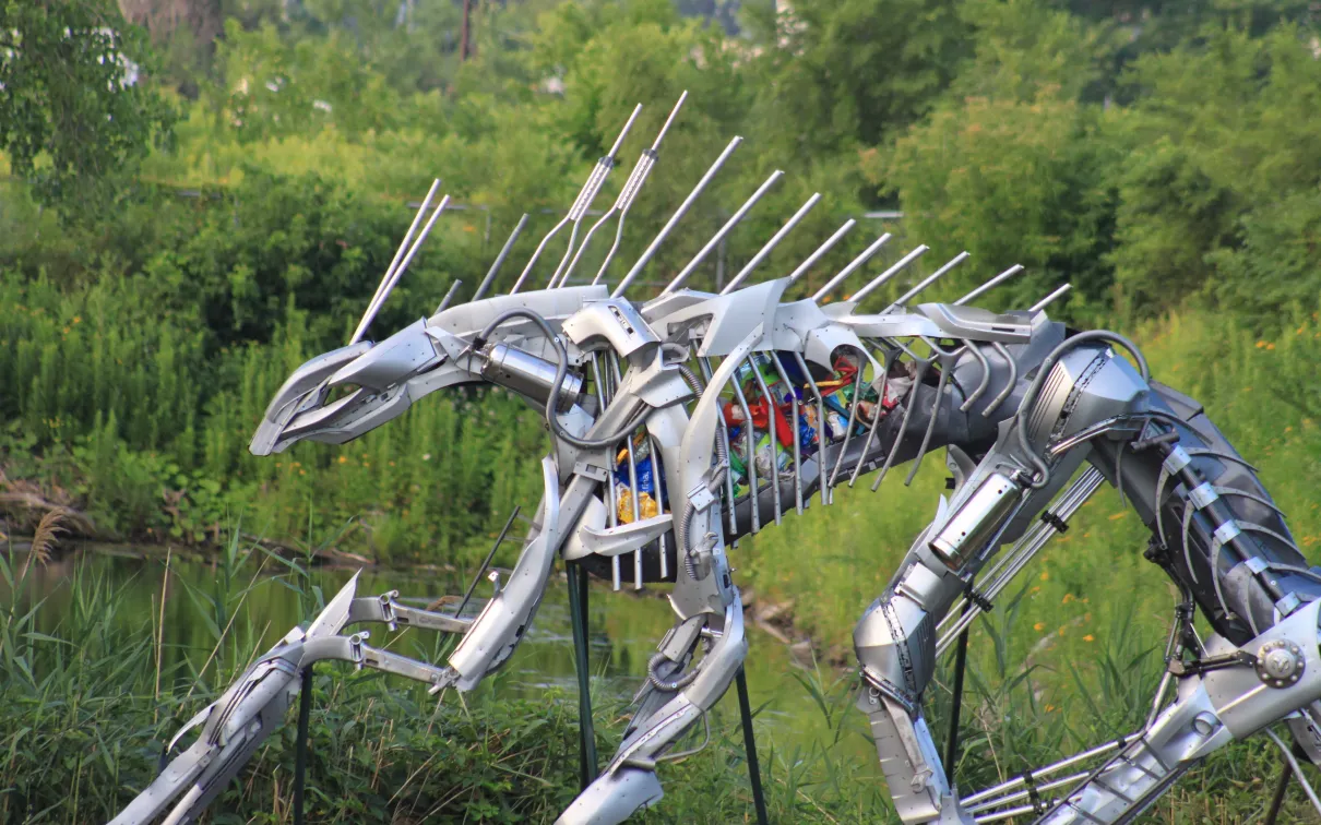 Large metallic sculpture of an insect with multiple legs and antennae, set outdoors with greenery in the background.