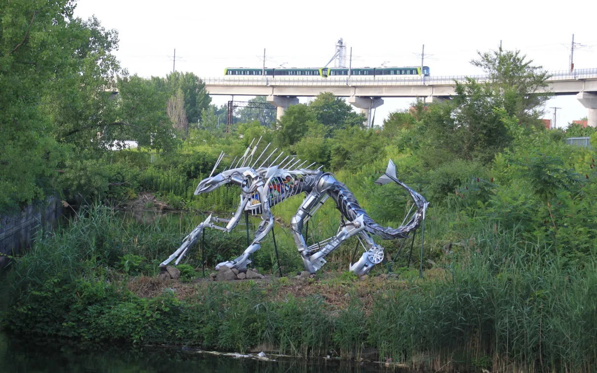 Steel sculpture of a crouching sabre-toothed cat surrounded by greenery near a body of water, with a passing train in the background.