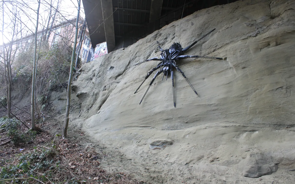 Large black spider sculpture mounted on a sandy rock cliff outdoors among bare trees.