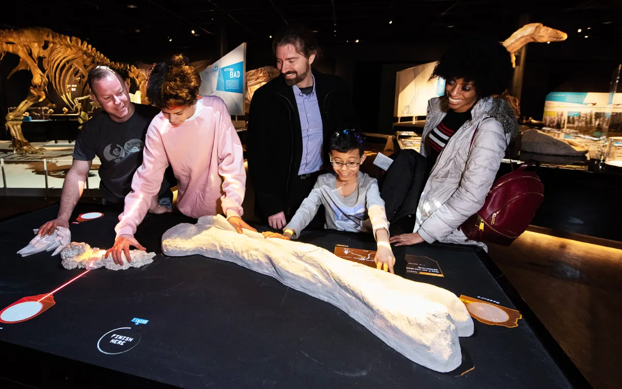 Five people, including a child, examine a large dinosaur bone displayed on a black table in a dimly lit museum gallery.