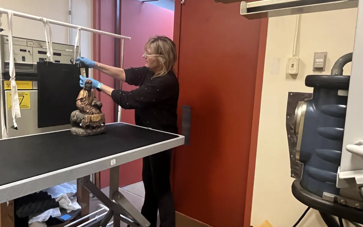 A woman adjusts a device near a Buddha statue on a metal table beside shelving and a red door.