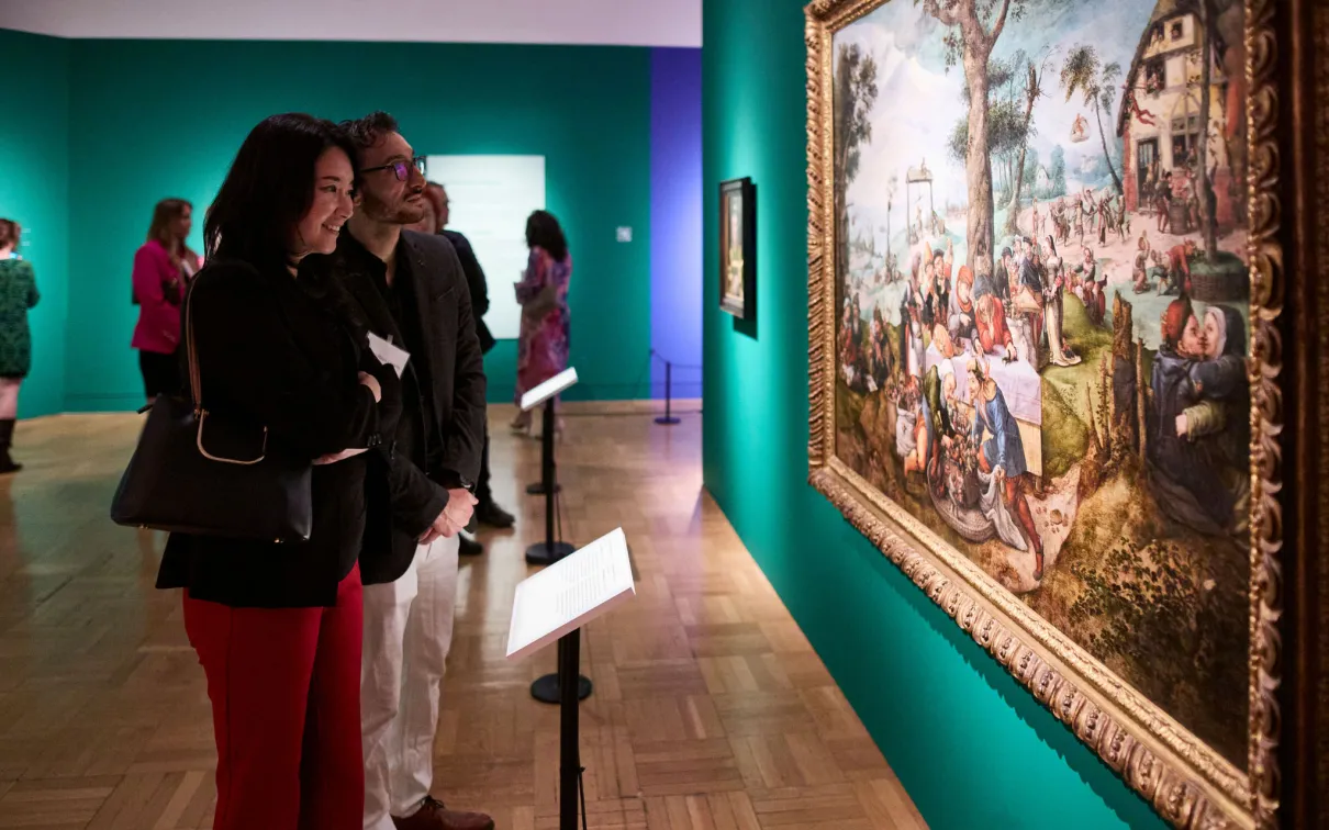 Two visitors closely observing a large framed painting displayed on a teal gallery wall.