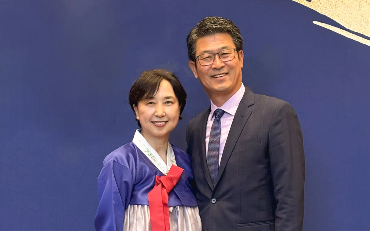 A man in a dark suit and glasses stands next to a woman wearing a traditional Korean hanbok dress with a blue top and red bow.