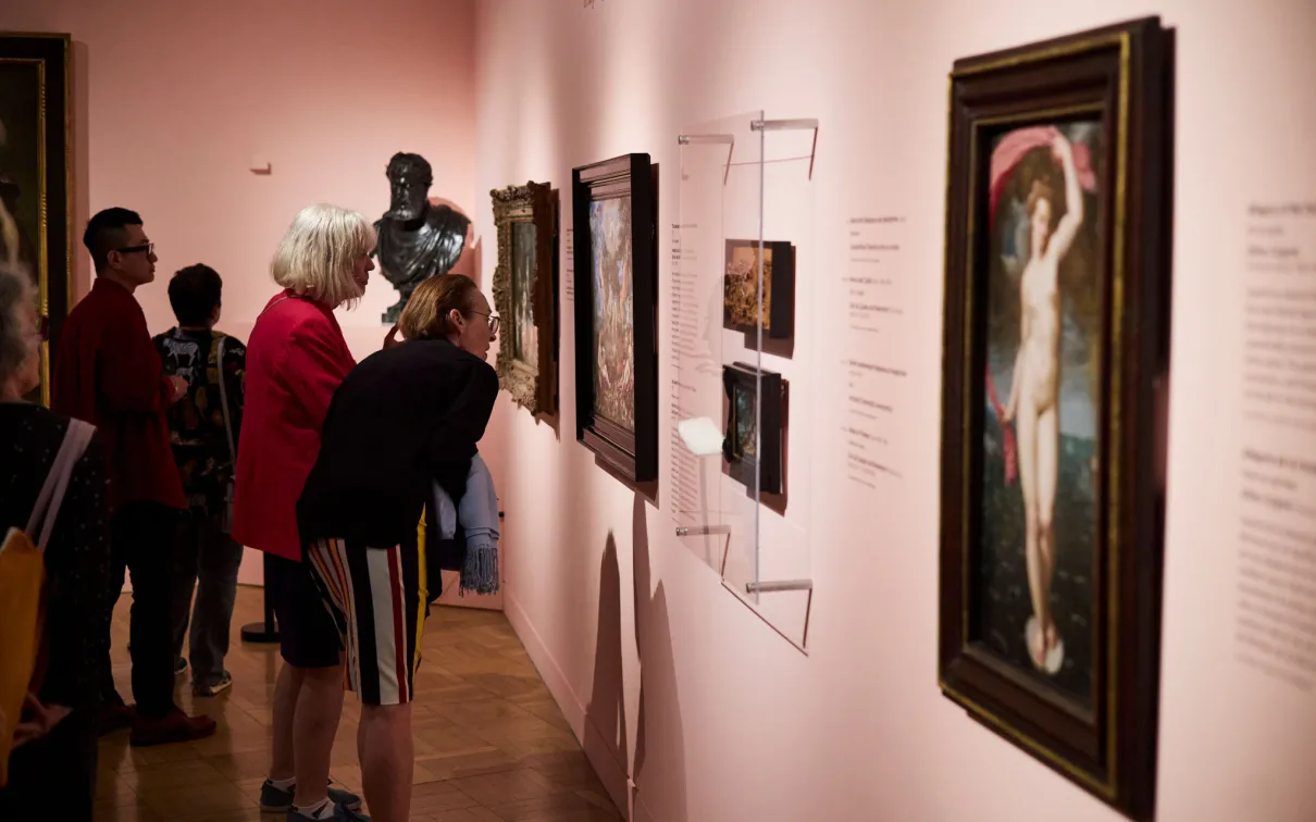 Several people closely observing framed artworks displayed on a museum wall with soft pink lighting.