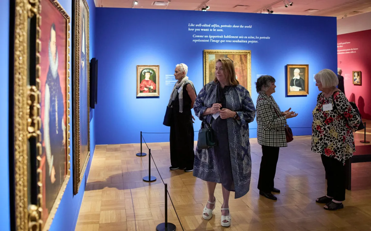 Five adults stand or walk inside an art gallery with blue and pink walls displaying framed paintings.