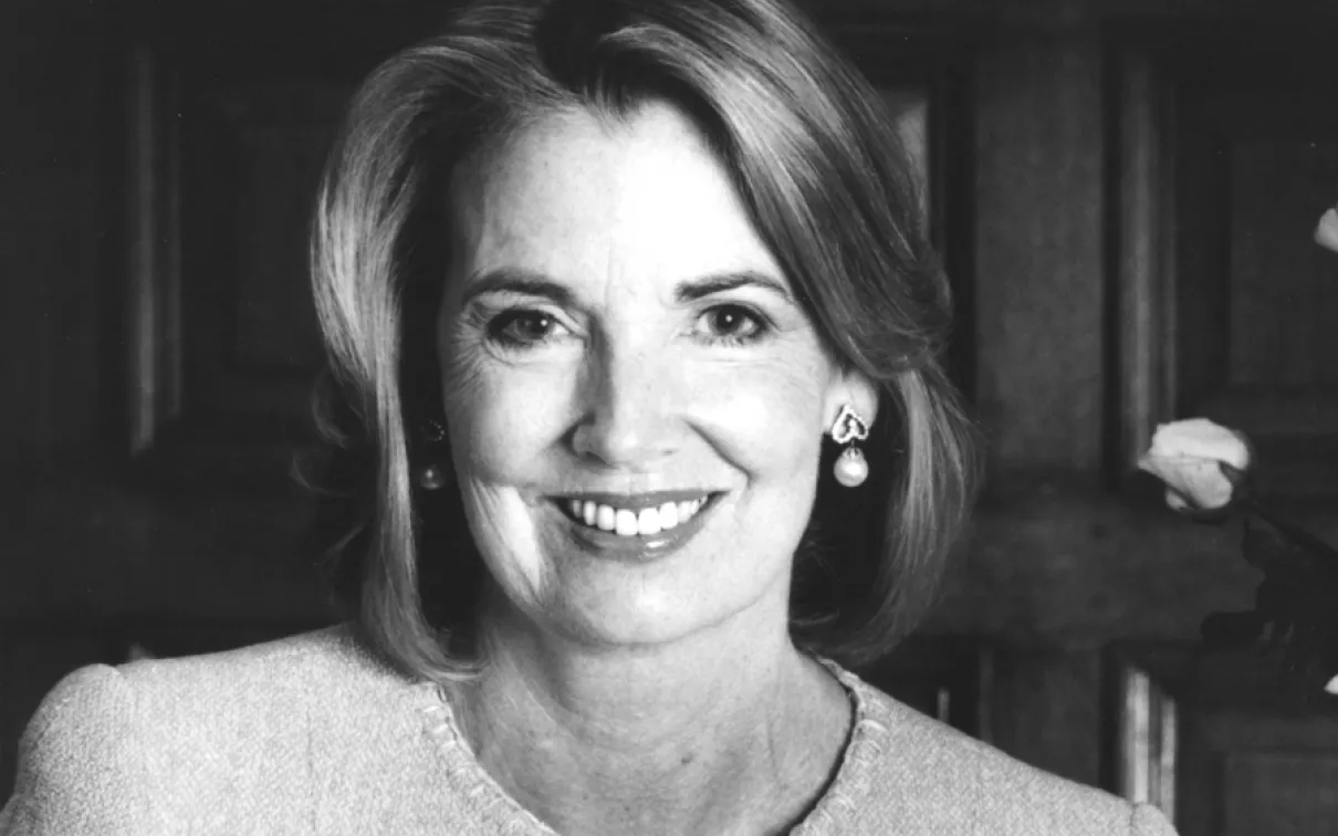 Portrait of a woman with light-colored hair, wearing a light jacket, smiling against a dark wooden background.