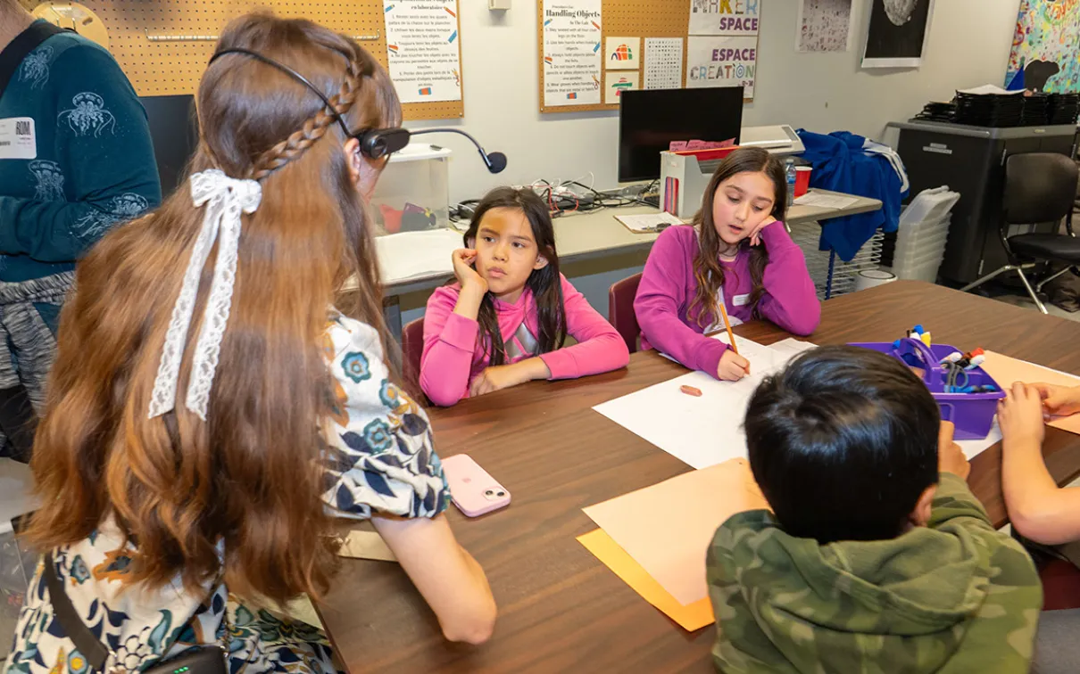 Students sitting at a table look up at a woman wearing a headset