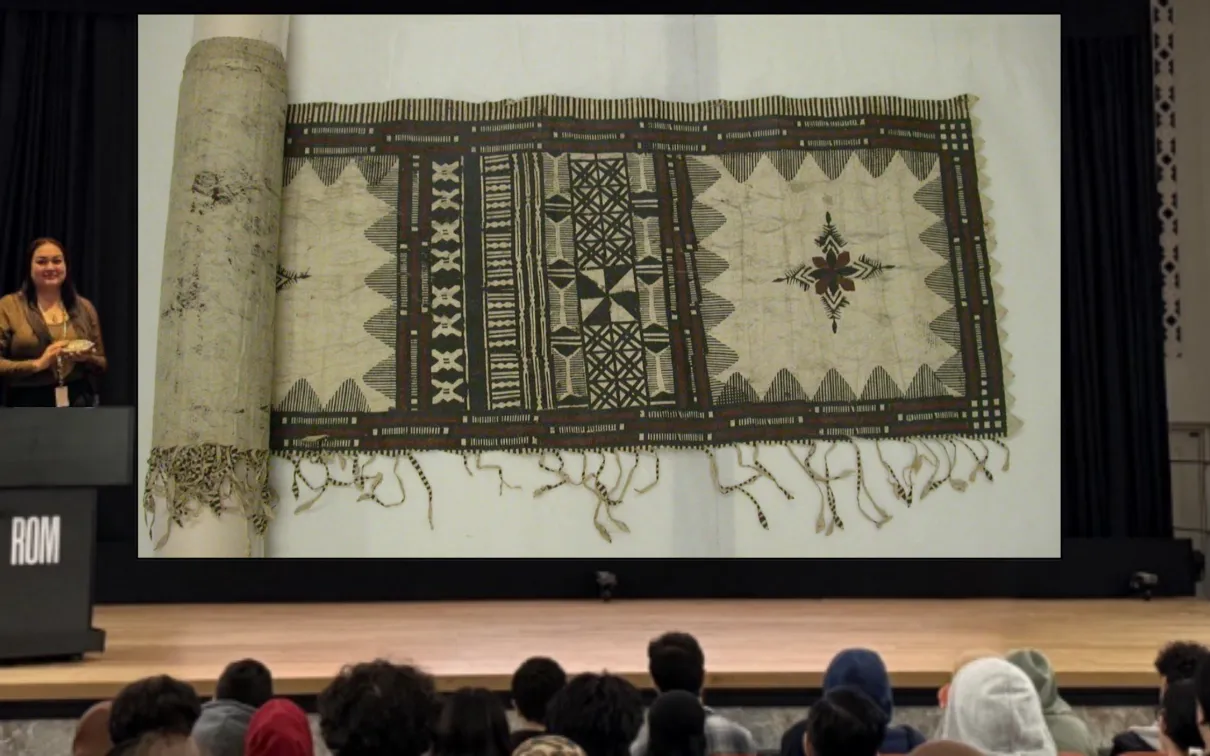 Une femme indigène souriante se tient derrière un podium sur une scène. Sur un écran à côté d'elle, on voit l'image d'un morceau de tissu avec des motifs complexes à l'encre noire et brune.