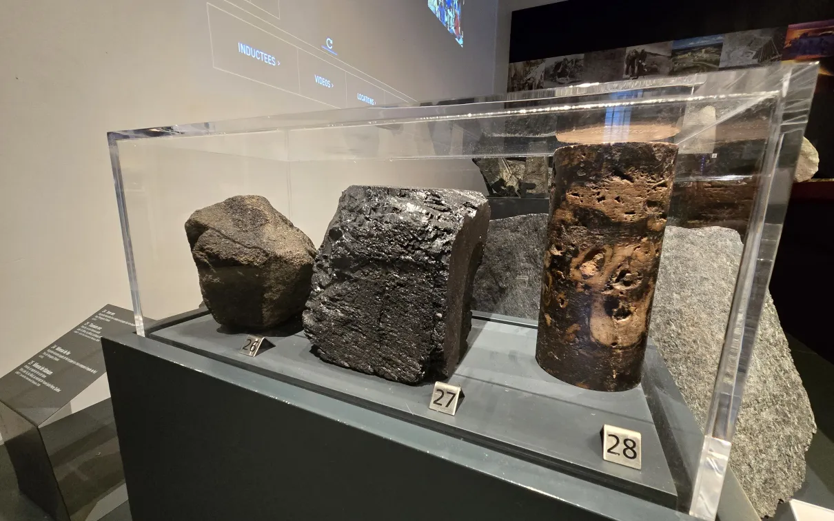 Dans la galerie d'un musée, une vitrine contient trois roches. L'une est brune/noire terne et arrondie, l'autre est noire et brillante, et la troisième présente des tons bruns avec des taches plus claires sur la roche plus foncée.