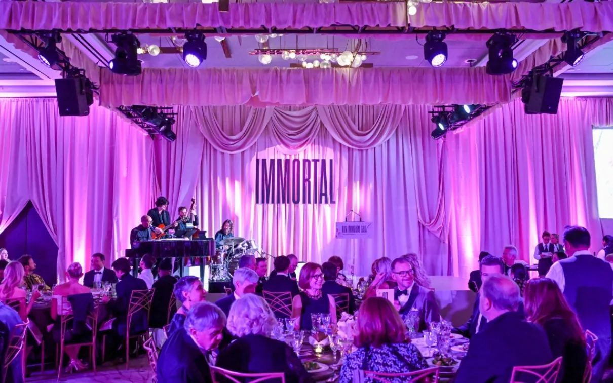 A banquet hall with round tables filled with people under purple lighting and a stage with musicians playing.