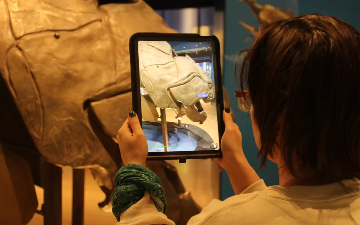 A person stands in front of a large fossil fish head holding an ipad that displays the head from the front.